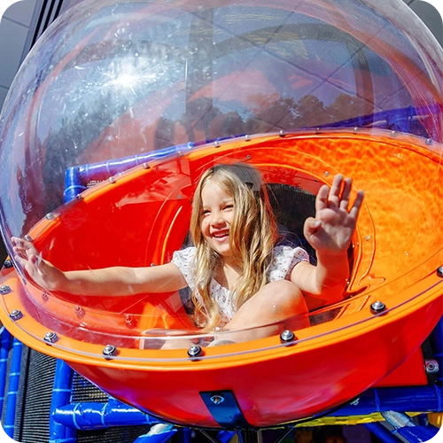 Smiling child playing inside a colorful enclosed tube slide at an indoor playground on a bright sunny day.